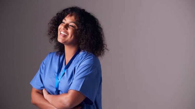 Studio Portrait Of Female Nurse Wearing Scrubs Standing Against Grey Background