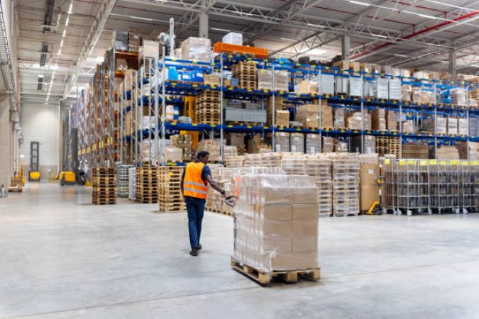 Rear view of a dock worker walking with packages on pallet jack at distribution warehouse. Young male dispatcher pulling pallet jack with boxes.