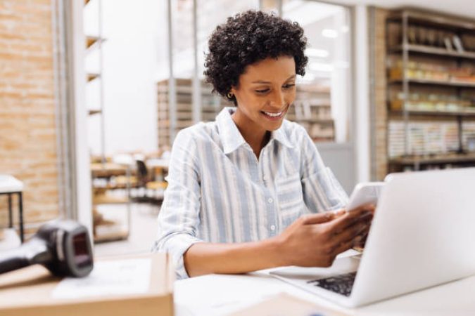 Cheerful online store manager reading a text message on her cellphone. Female entrepreneur making plans for product shipping in her warehouse. Creative businesswoman running an e-commerce small business.